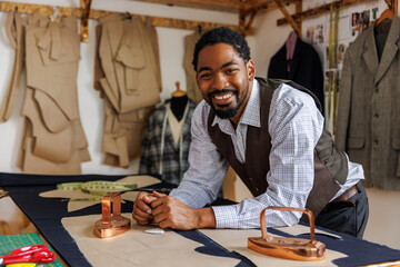 Portrait of smiling tailor standing at table with tools in workshop