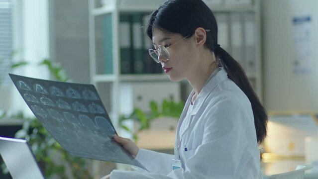 Young Asian female doctor examining CT scan and typing on laptop when working in clinic