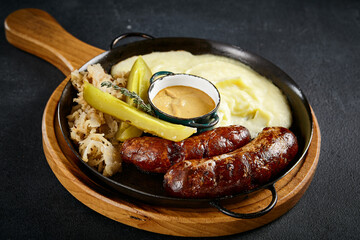Grilled sausage with mashed potatoes and pickles presented on a wooden board on a dark background. Classic comfort food with a rustic touch