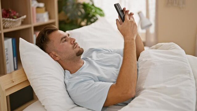A young hispanic man with a beard uses his smartphone in bed, suggesting restlessness before sleep in a comfortable home setting.