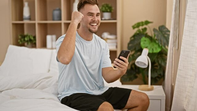 A cheerful young man in casual wear using a smartphone in his modern bedroom communicates excitement and joy.