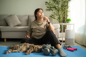 Happy young overweight obese woman resting on fitness mat and drinking fresh water from bottle.