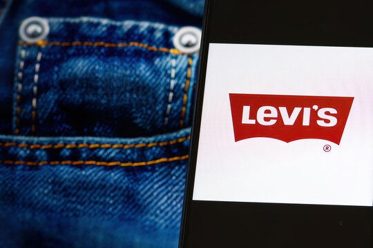 Levi's company logo on screen of smartphone against blurred background with blue jeans image displayed on computer screen.