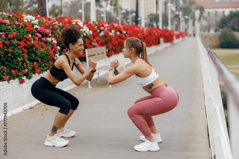 Wall mural two active young women doing workout and squatting together outdoors in the city