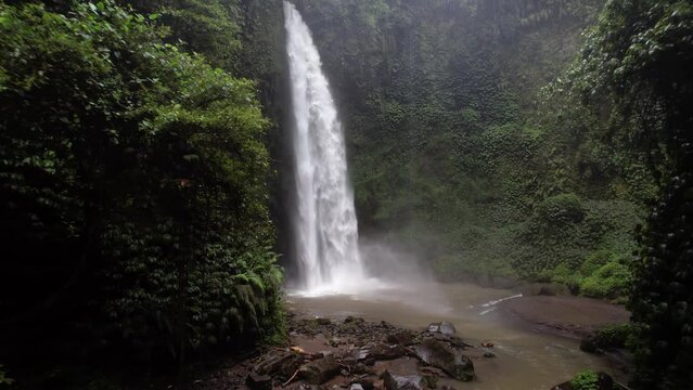 Big waterfall in green canyon, camera fly back, short clip of Nungnung. Water fall down from height to shallow pond and splash, white water mist in air around. Tropical plants covering vertical walls