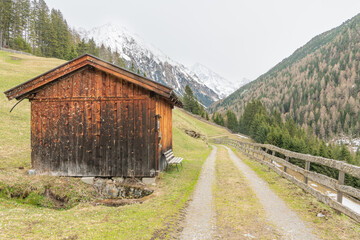 Hiking path between grassy alpine slopes viewing towards pine trees and mountains in Gries,  during early spring time. Last snow on the trail. Besinnungsweg trail in Gries, Längenfeld.Wooden barn.