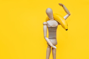 A wooden mannequin of a man holds a banana in his hands on a yellow background. The concept of healthy nutrition, fruit diet and high-calorie foods.