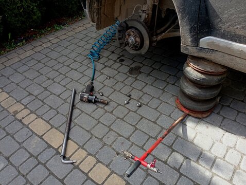 The front of the car is raised on a lift and with the wheel removed. There is a pneumatic wrench near the car and the bolts that hold the car wheel are lying on the cobblestones. The topic of seasonal