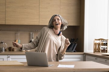 Happy mature woman enjoy track, dancing, stand at kitchen table with laptop, distracted from work,...
