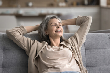 Close up of smiling middle-aged woman relaxing on cozy sofa with eyes closed, put hands behind...