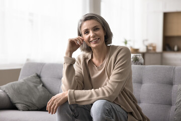 Cheerful mature woman relaxing on sofa, look at camera having appealing appearance, perfect white...