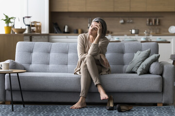 Drained mature woman came home after long stressful workday took off her shoes, resting seated on sofa, feels unwell, squeezed like lemon, having problems, suffer from lack of sleep, chronic fatigue - obrazy, fototapety, plakaty