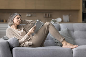 Pretty mature woman resting on comfortable sofa with book, feel satisfied reading interesting literature, favorite novel or poetry, spend weekend time alone indoors, booklover, hobby, pastime at home
