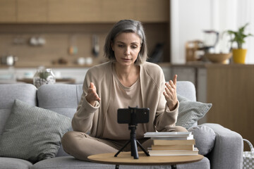 Middle aged woman sitting on sofa in living room records new videoblog, speak at cellphone camera fixed on tripod, share experience, talk to internet audience. Online communication, coaching, blogging