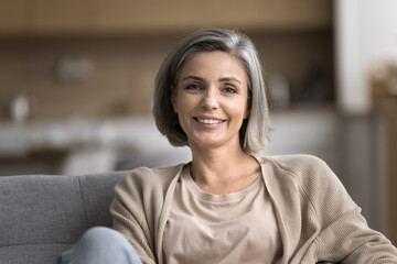 Close up portrait of smiling mature woman looking at camera, pose for photo, relax on cozy couch in living room, feels carefree on weekend alone indoors, pleasant happy lady enjoy leisure time at home