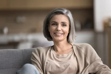 Poster Im Rahmen Close up portrait of smiling mature woman looking at camera, pose for photo, relax on cozy couch in living room, feels carefree on weekend alone indoors, pleasant happy lady enjoy leisure time at home © fizkes