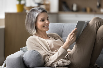 Carefree happy middle-aged woman spend time at home, read interesting book, lies on comfortable...