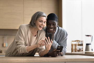 Laughing interracial couple spend time at home, stand at kitchen table with two smart phones, have...