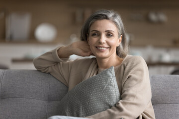 Beautiful pensive middle-aged woman embrace cushion enjoy weekend relaxation seated on comfortable couch in apartment. Carefree leisure at home, pleasant thoughts, optimistic mood, life satisfaction - obrazy, fototapety, plakaty