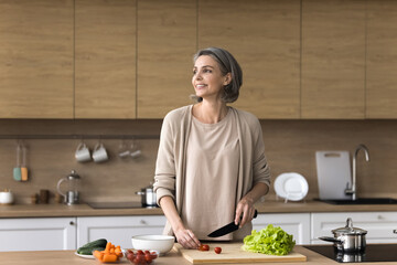 Smiling mature dreamy woman stand at table, cutting fresh vegetables for salad, looking away, smile, planning dinner menu, use healthy ingredients, enjoy cooking process in modern kitchen. Culinary - 778022706
