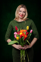 A beautiful middle-aged blonde woman holds a bouquet of tulip flowers, smiles and looks at the camera on a dark background.