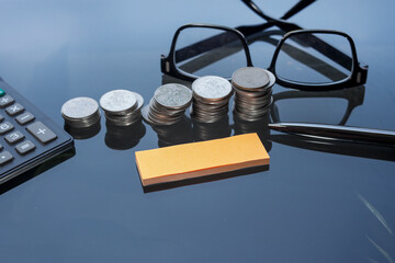 Empty post-note, stacked coins, pen, eyeglasses, and calculator on the glass table. Work space and...