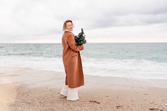 Blond woman Christmas tree sea. Christmas portrait of a happy woman walking along the beach and holding a Christmas tree in her hands. She is wearing a brown coat and a white suit.