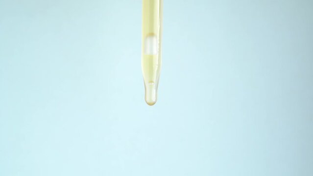 Close-up glass dropper with cosmetic serum on a blue background. Essential oil, body serum, drops drip. Drops of serum or essential oil fall from a glass pipette.