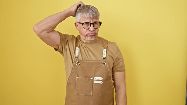 Uncertain grey-haired man in glasses and apron, pensive and full of doubt, standing alone on yellow background, wondering and thinking about a confusing question.