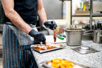 A chef meticulously applies sauce to grilled meat in a professional kitchen, surrounded by utensils...