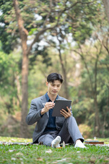 Portrait of businessman sitting on green grass in the city park and using digital tablet