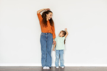 Young mother and her little girl measuring their heights near light wall indoors, looking at each other and smiling, full length shot
