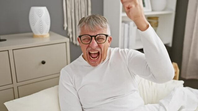Furious, aggressive middle-aged grey-haired man in pyjamas, frantic with anger, raises fist in bedroom
