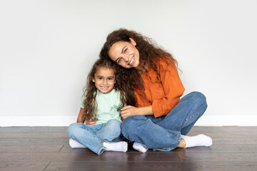 Obraz na płótnie Canvas Happy mom and female child bonding together, sitting on floor over white wall at home, mother and daughter embracing and smiling