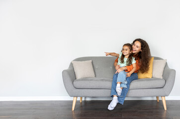 Mom and cute daughter pointing aside at free space while relaxing on sofa over white wall, showing...
