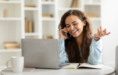 Positive freelancer woman enjoying job communication, sitting at home, looking at laptop and talking on phone, smiling