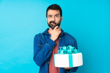 Young handsome man with a big cake over isolated blue background having doubts and with confuse...