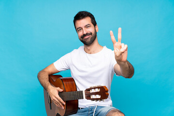 Young man with guitar over isolated blue background smiling and showing victory sign