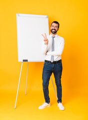 Full-length shot of businessman giving a presentation on white board over isolated yellow background smiling and showing victory sign