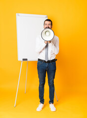 Full-length shot of businessman giving a presentation on white board over isolated yellow background shouting through a megaphone - 778099136