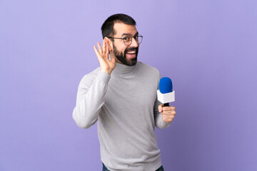 Adult reporter man with beard holding a microphone over isolated purple background listening to...