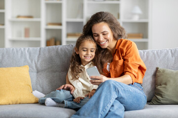 Cheerful mother and daughter using smartphone together, sitting on couch at home, shopping online...