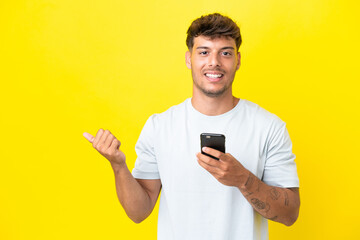 Young caucasian handsome man isolated on yellow background using mobile phone and pointing to the lateral