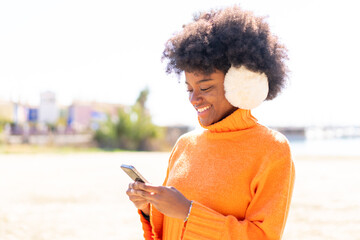 African American girl wearing winter muffs at outdoors sending a message with the mobile