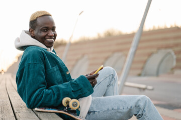 Smiling young skater man in a modern city. Afro American guy portrait with mobile phone.