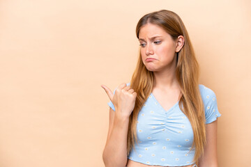 Young caucasian woman isolated on beige background unhappy and pointing to the side