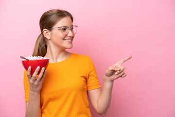 Young caucasian woman holding a bowl of cereals isolated on pink background pointing to the side to...