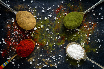 Spoons with spices and salt on black background sprinkled with different kind of spices - obrazy, fototapety, plakaty