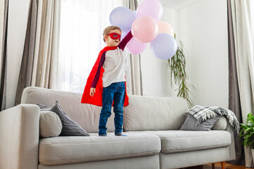 Young child in superhero costume holding balloons on a couch
