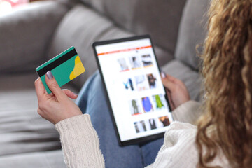 Back close-up of credit card together with white girl using an online store on the sofa with a...